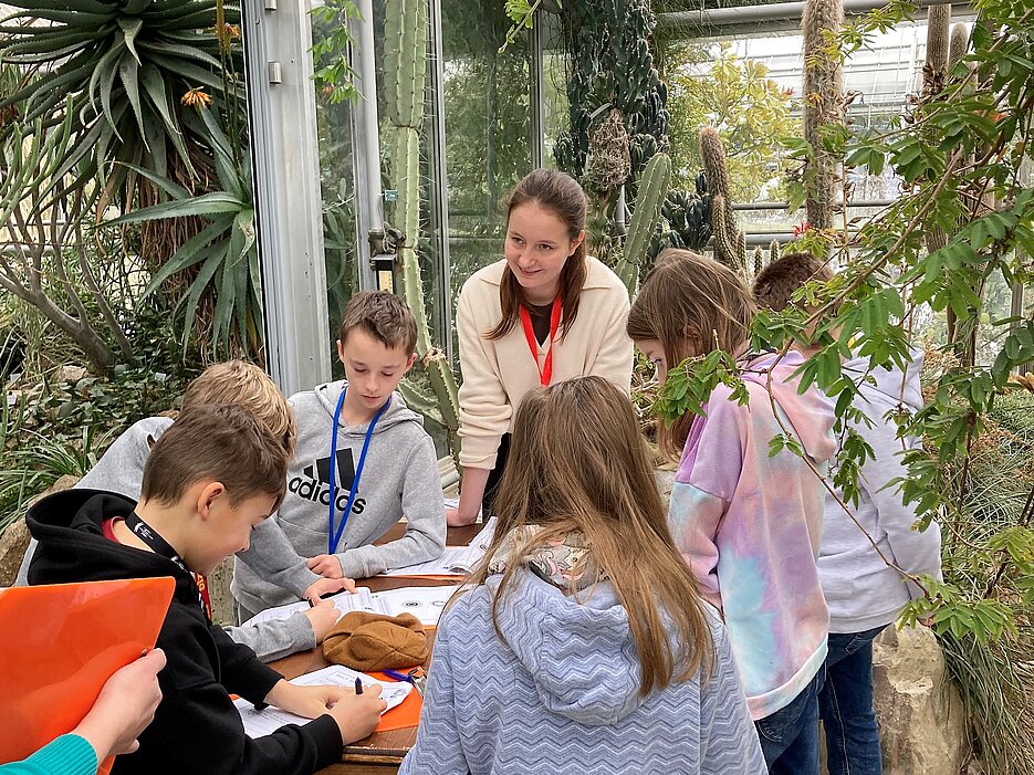 Das Foto zeigt eine Gruppe von Schüler:innen einer Grundschule mit einer Studentin bei der Stationenarbeit im Tropenschauhaus des Botanischen Gartens.