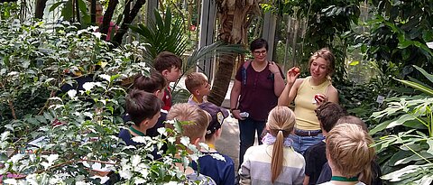 Das Foto zeigt eine Studentin mit einer Gruppe von Schüler:innen an der Workshop-Station zu saisonalen und regionalen Früchten im tropischen Nutzpflanzenhaus des Botanischen Gartens.
